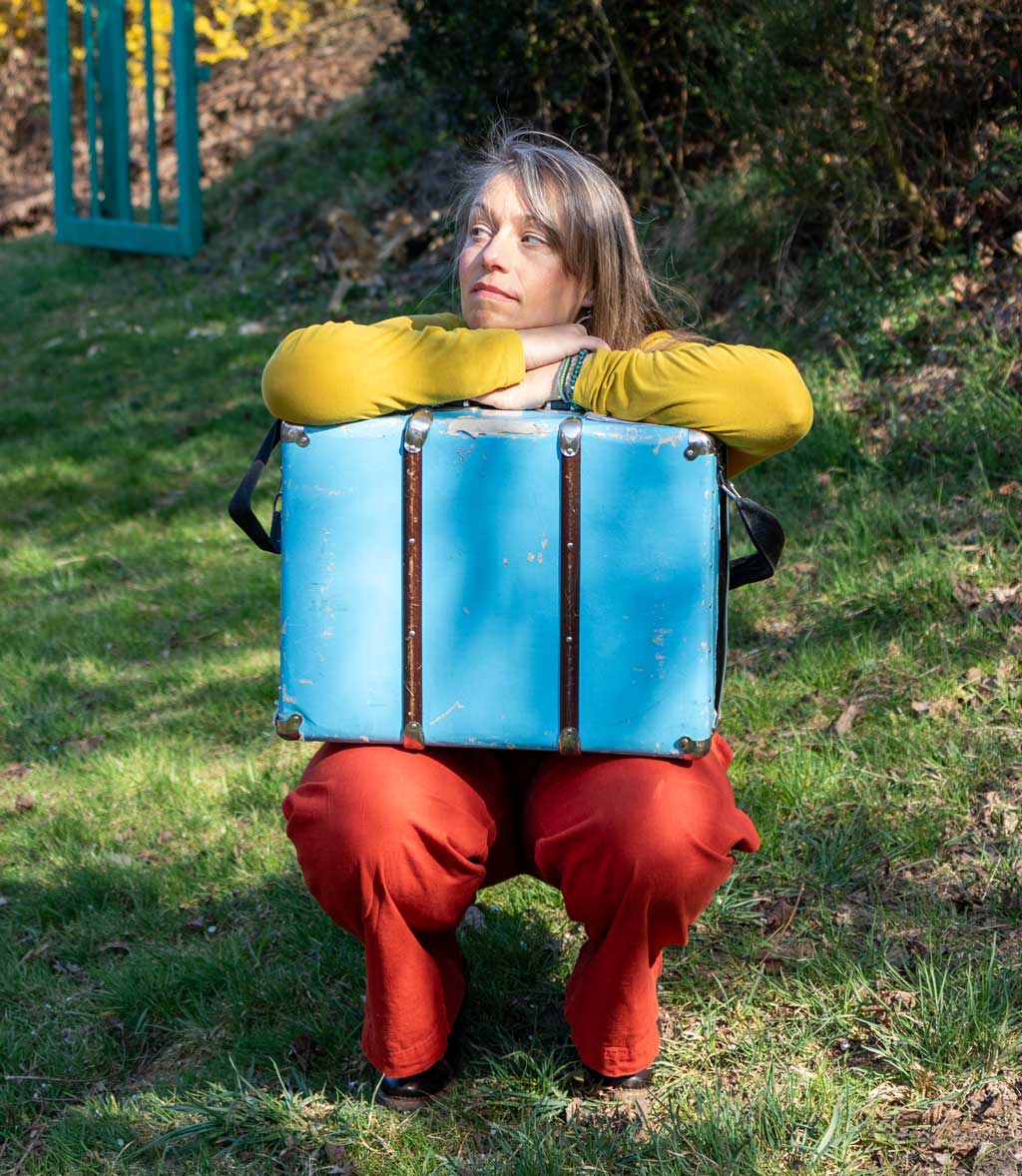 Anna Stijohann mit einem Koffer auf einer Wiese, im freundlichen Frühlingslicht