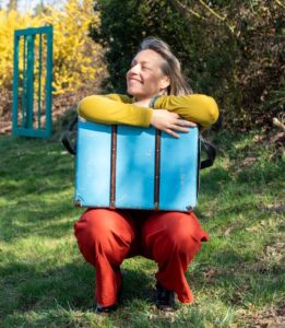 Anna Stijohann mit einem Koffer auf einer Wiese, im freundlichen Frühlingslicht