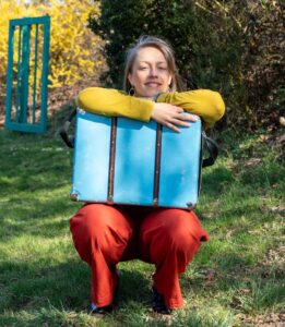 Anna Stijohann mit einem Koffer auf einer Wiese, im freundlichen Frühlingslicht